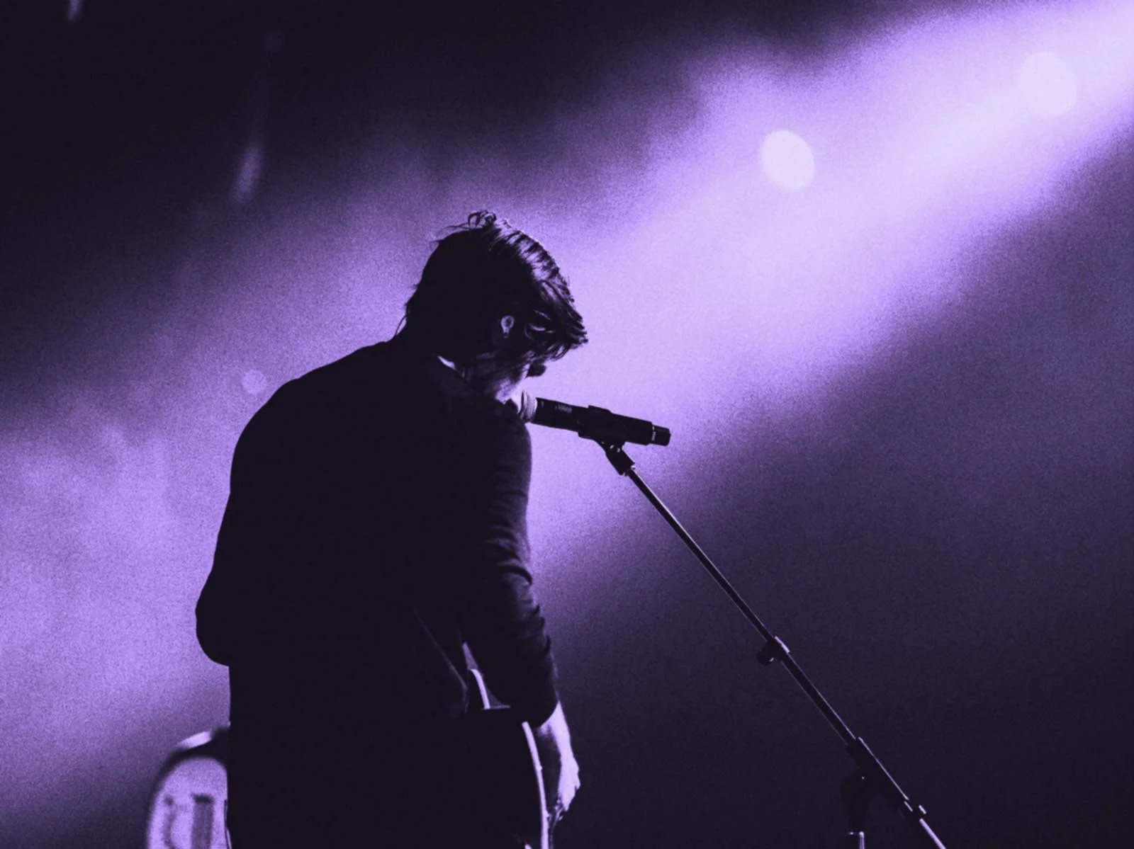 Silhouette of a male musician with long hair standing near a microphone on stage, illuminated by purple stage lights.