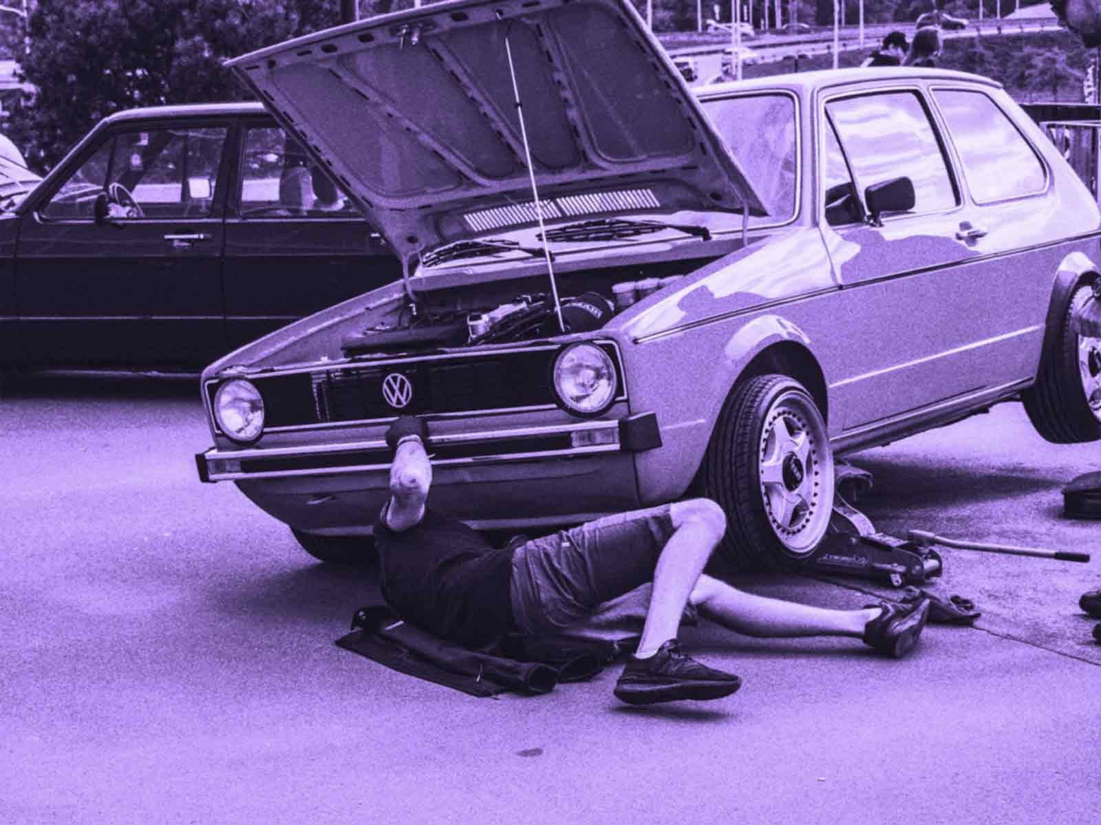 A person lying on the ground working underneath a vintage Volkswagen car with the hood open, on a concrete surface outdoors.