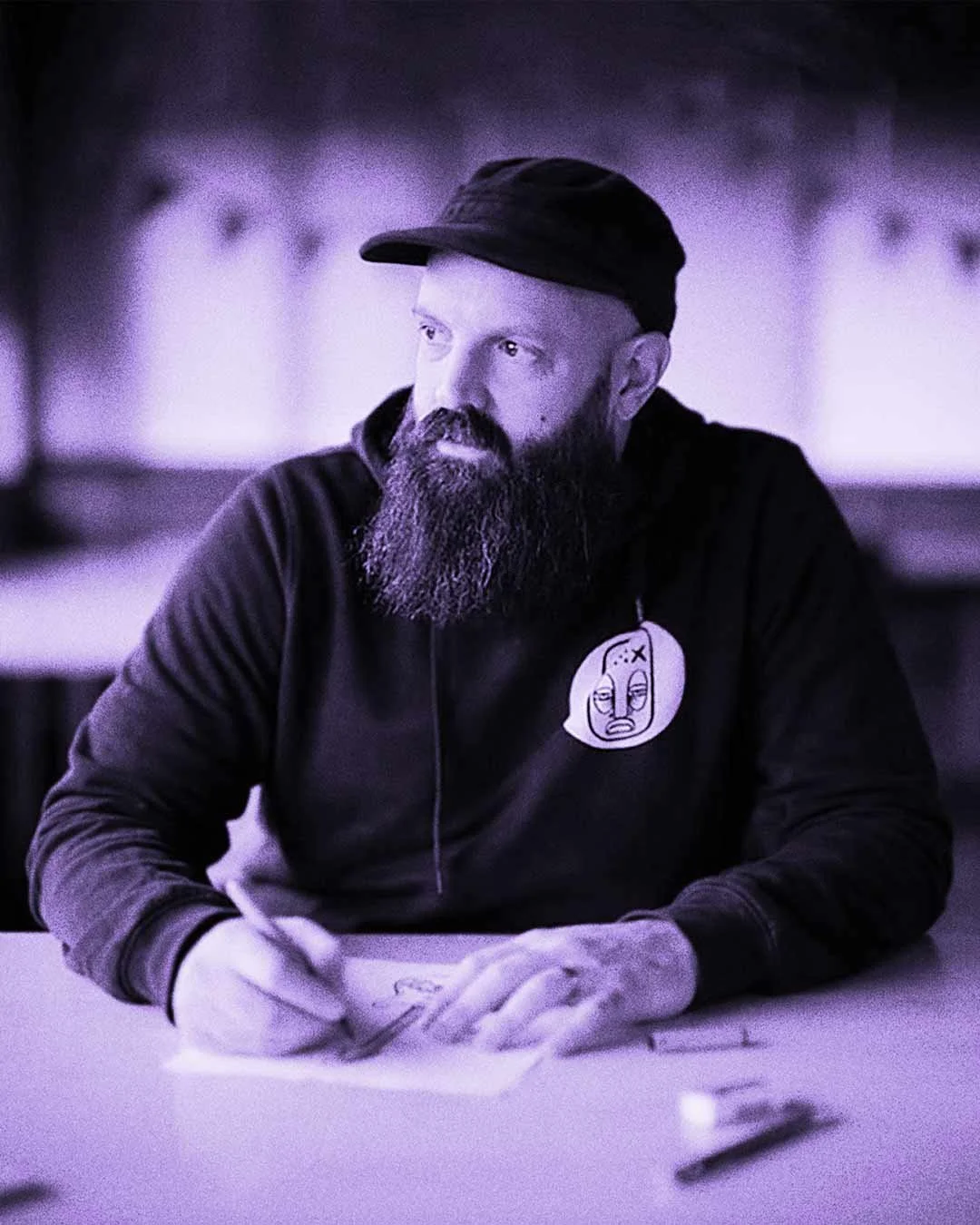 A bearded man in a baseball cap draws on a piece of paper.