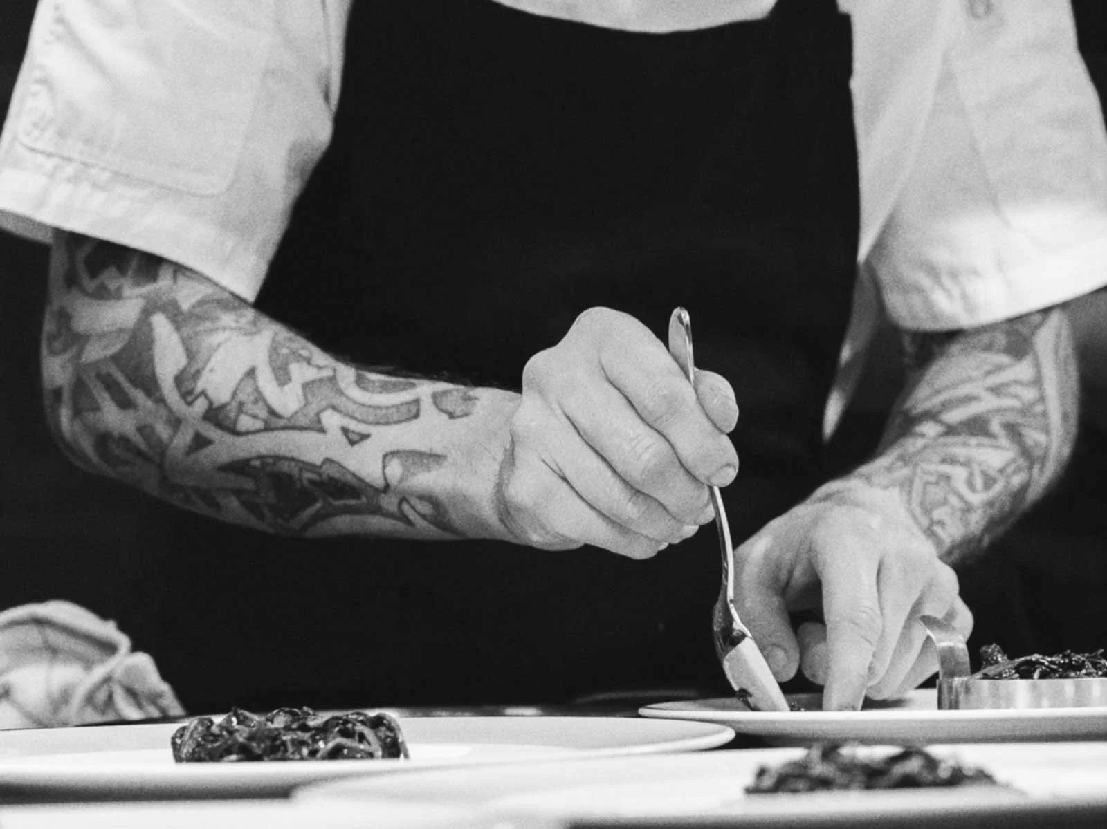 A chef with tattooed arms, wearing a black apron and white shirt, holding a spoon and serving food on a plate, with other plates of food in the foreground.