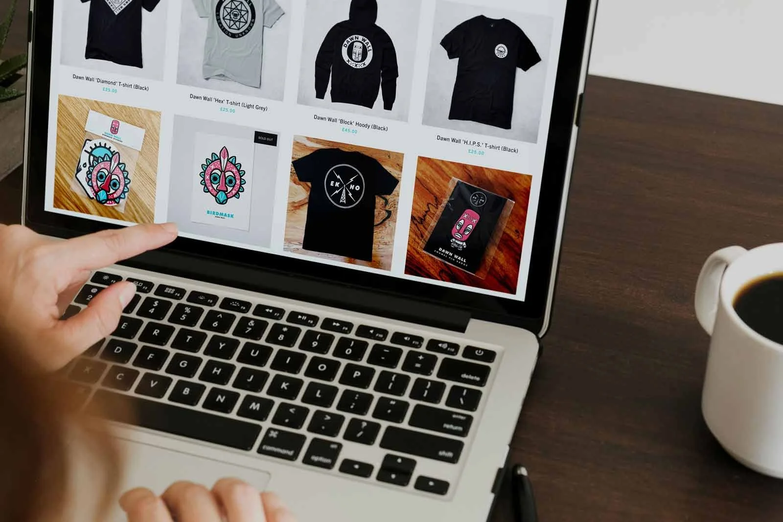 A hand points at a laptop displaying an online store with t-shirts and stickers.