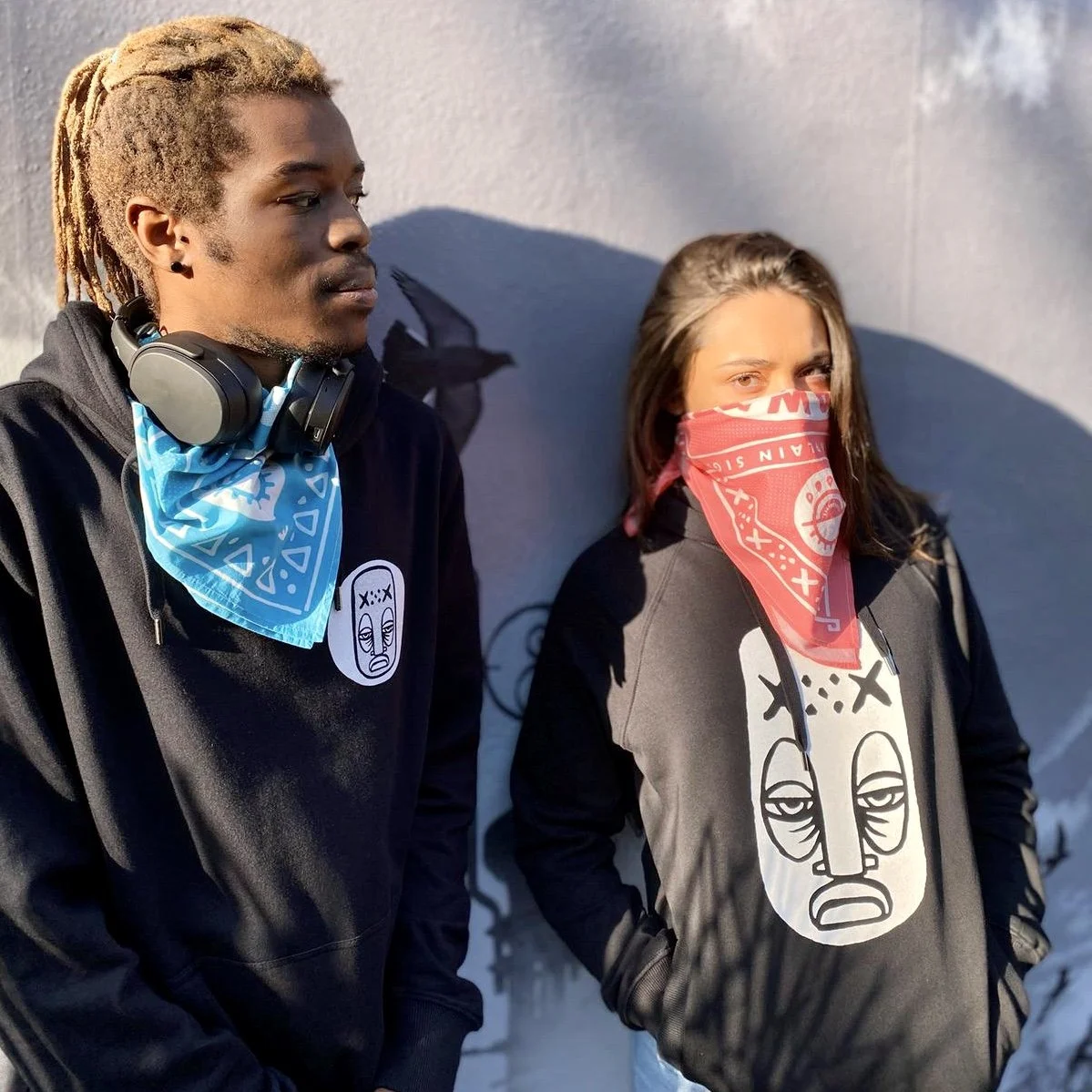 Two people in black hoodies with graphic masks and bandanas stand against a wall.