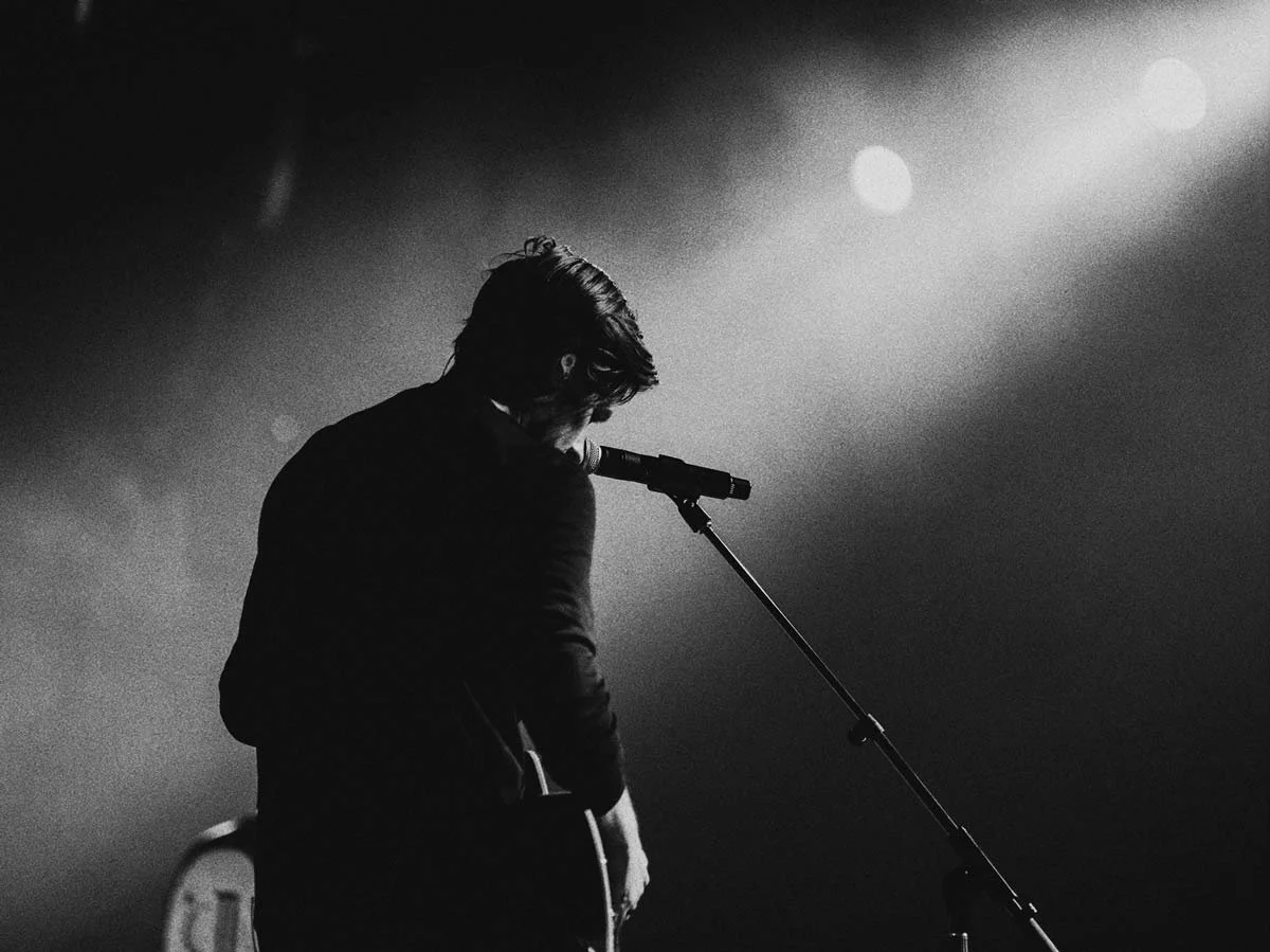 A black and white photo of a man holding a guitar and standing in front of a microphone on a dimly lit stage.