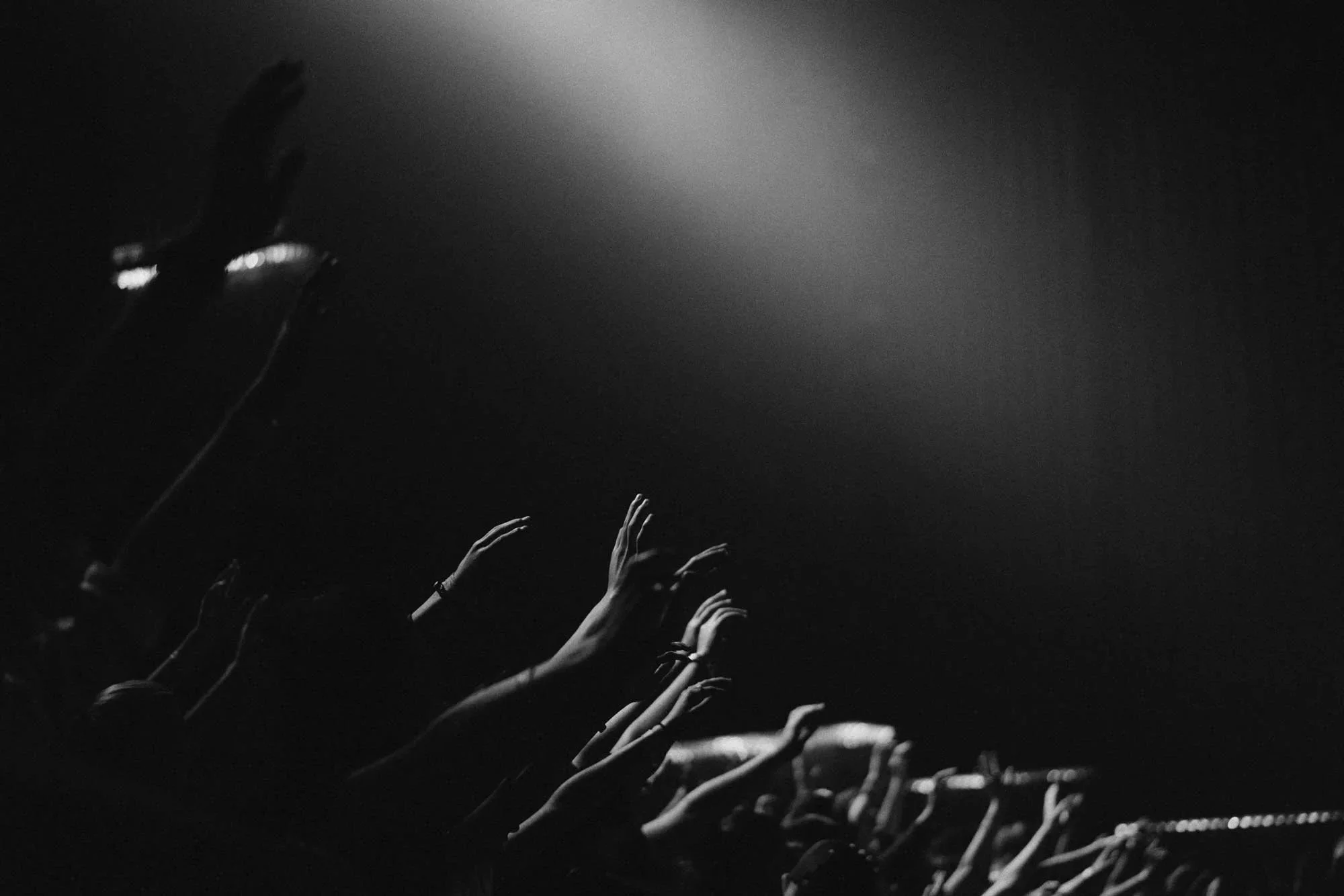 Black and white photo of concert audience with raised hands reaching towards a bright light shining from above.