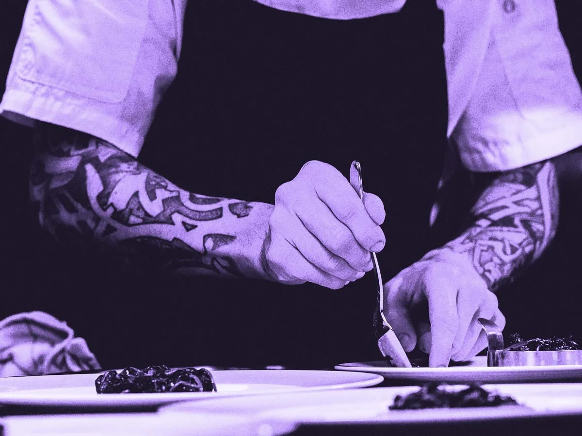 Chef with tattooed arms plates food with a spoon on a white plate.