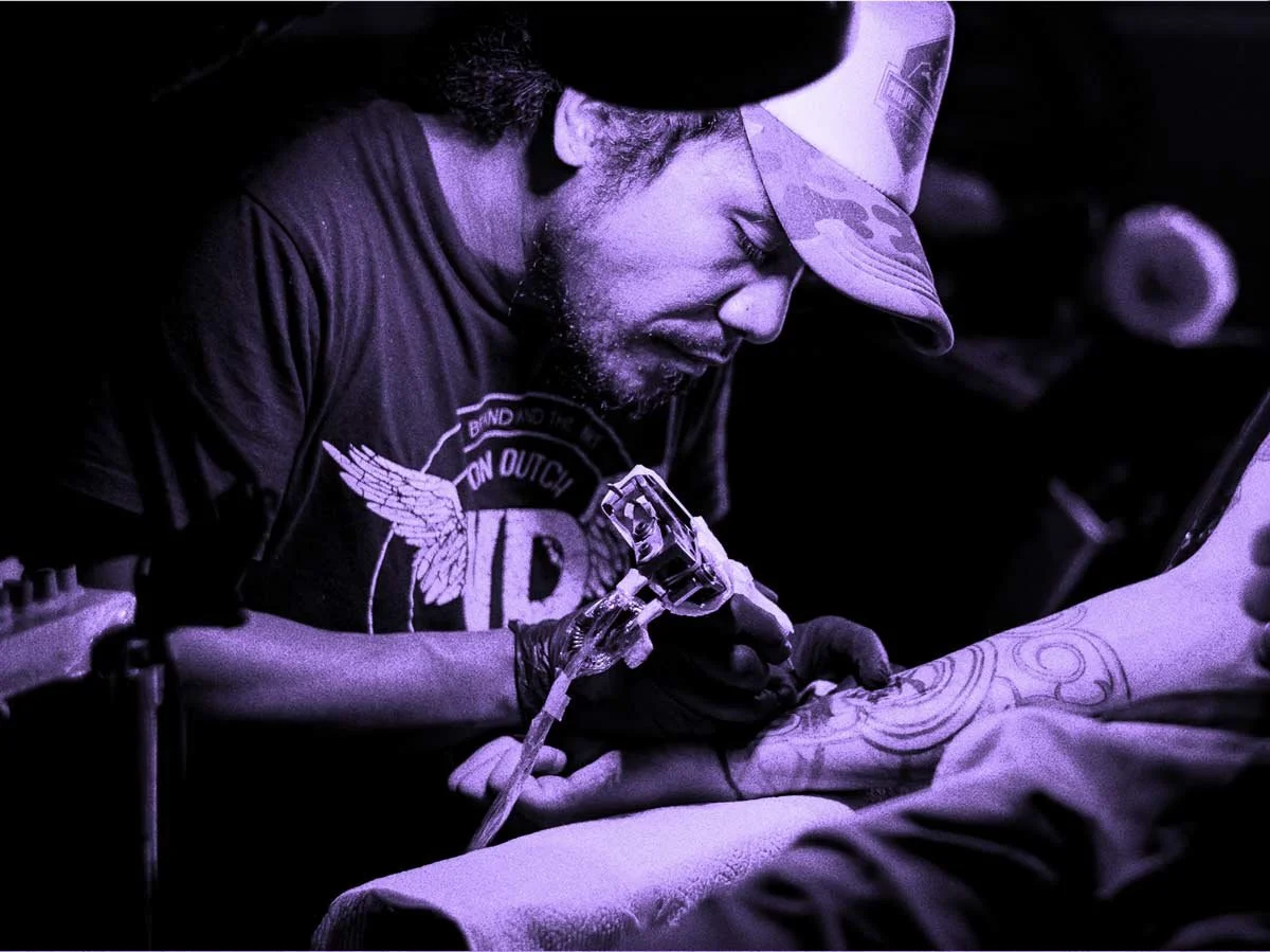 Tattoo artist with a beard and cap inks a design onto an arm.