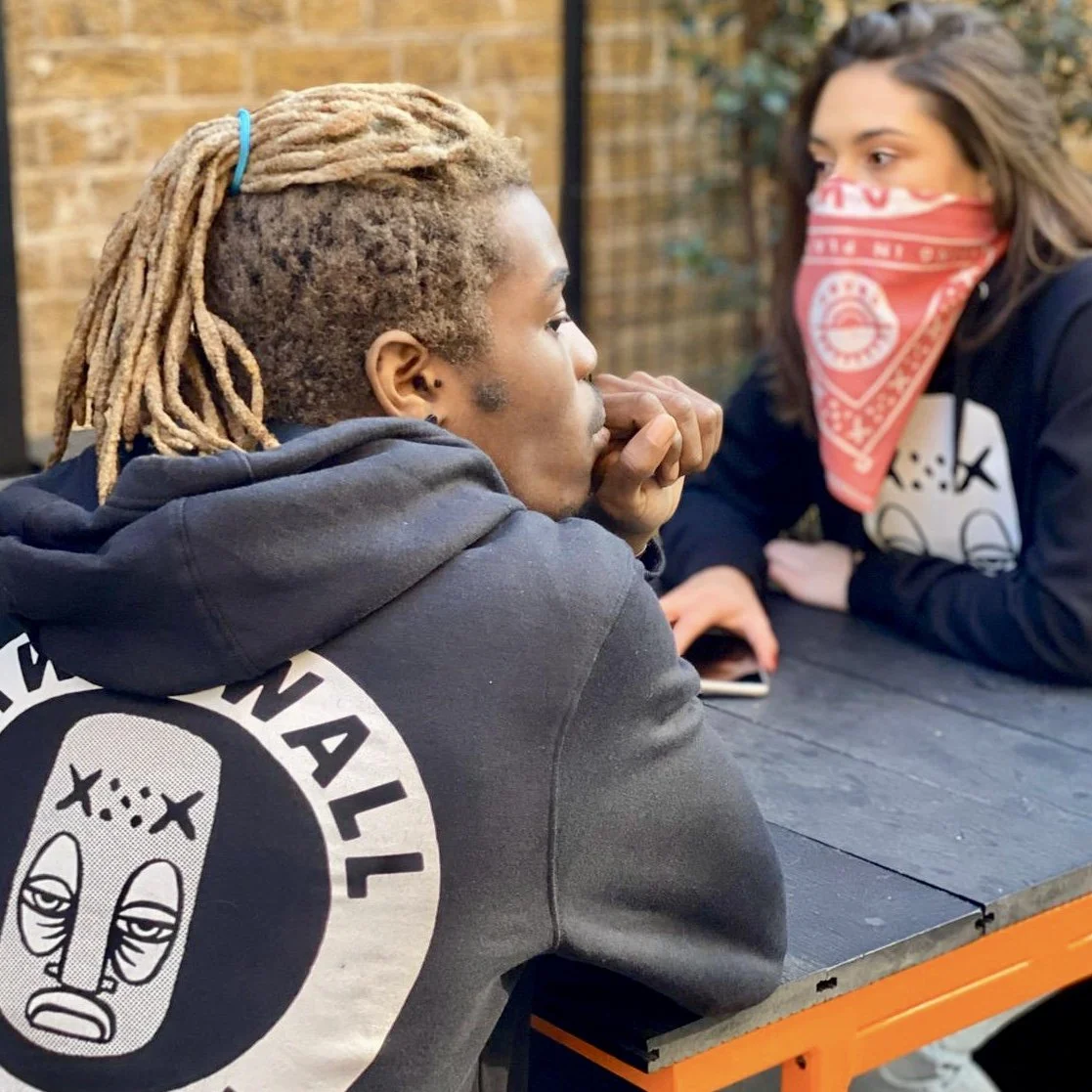 Two people sit at a table, one wearing a black hoodie with a cartoon face design.