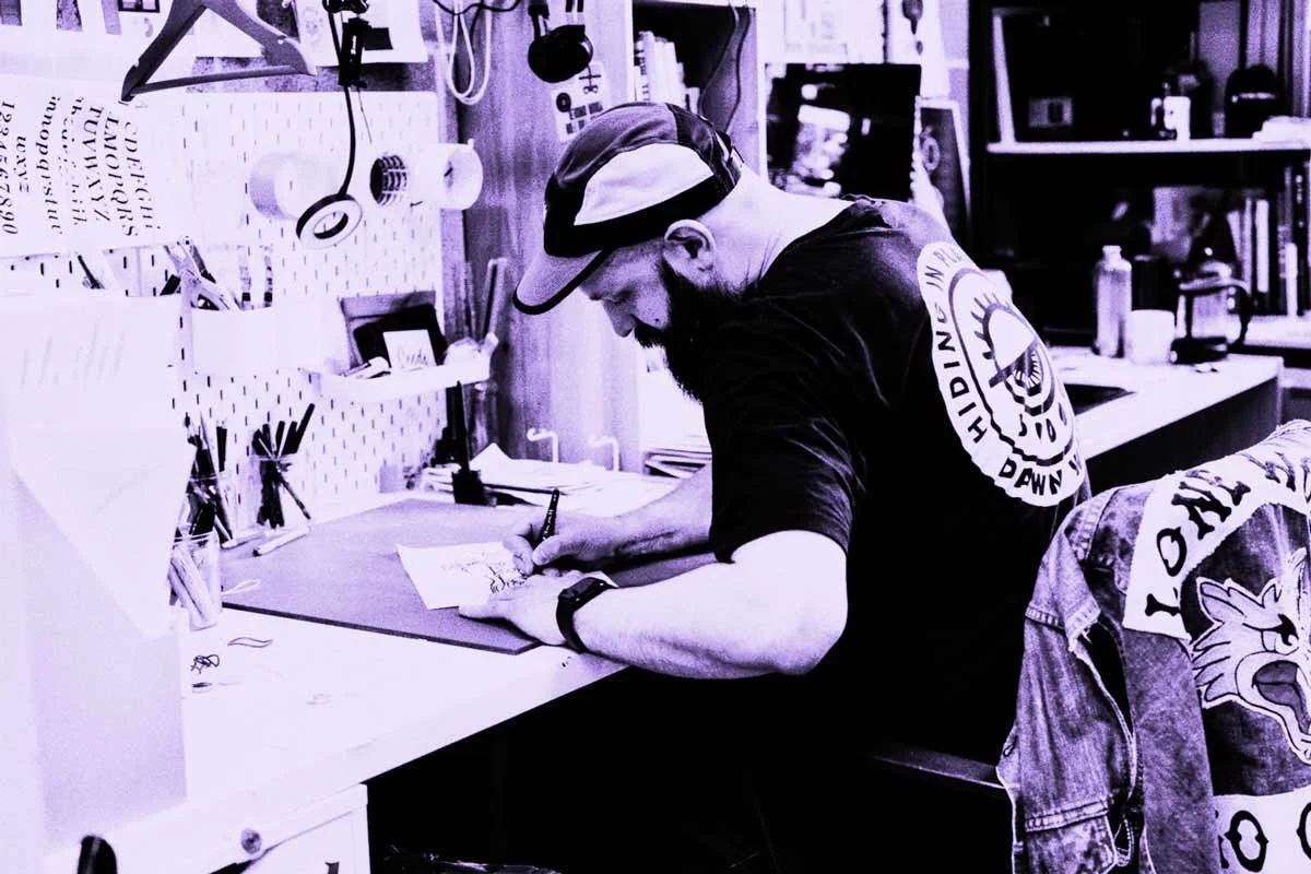Designer at a desk, drawing with a pen on paper, wearing a baseball cap and a t-shirt.