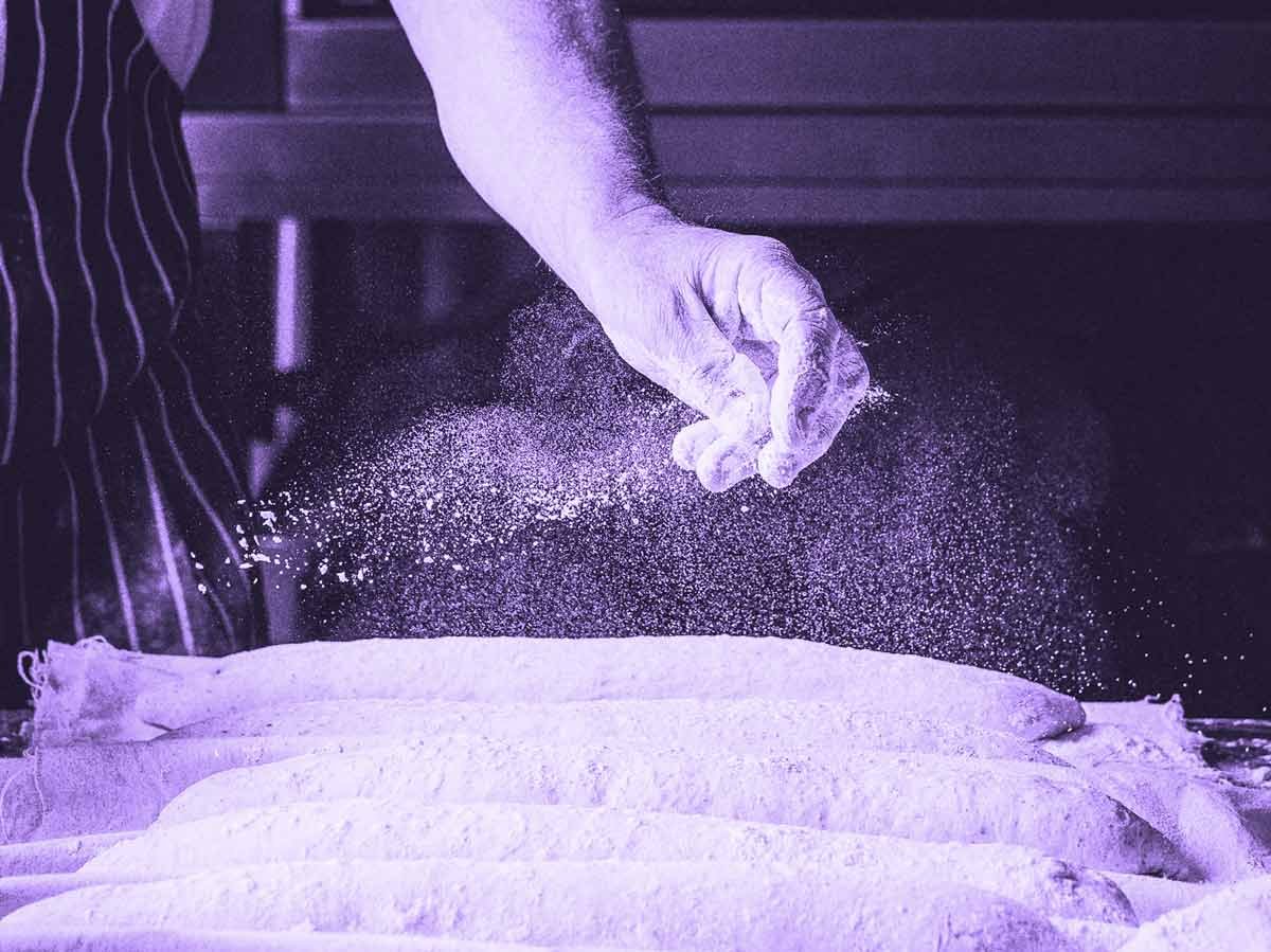 A baker's hand sprinkles flour over uncooked loaves of bread.