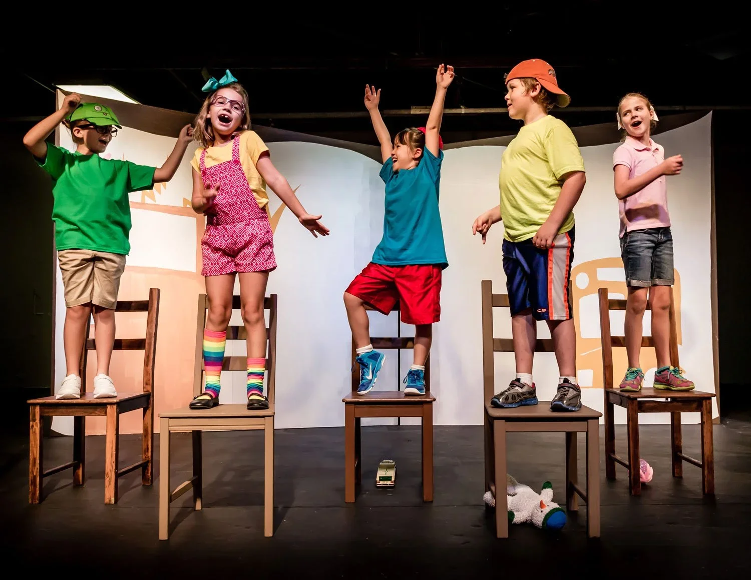 Group of five children on stage during a play or performance, standing on chairs with some smiling, laughing, or raising their arms.