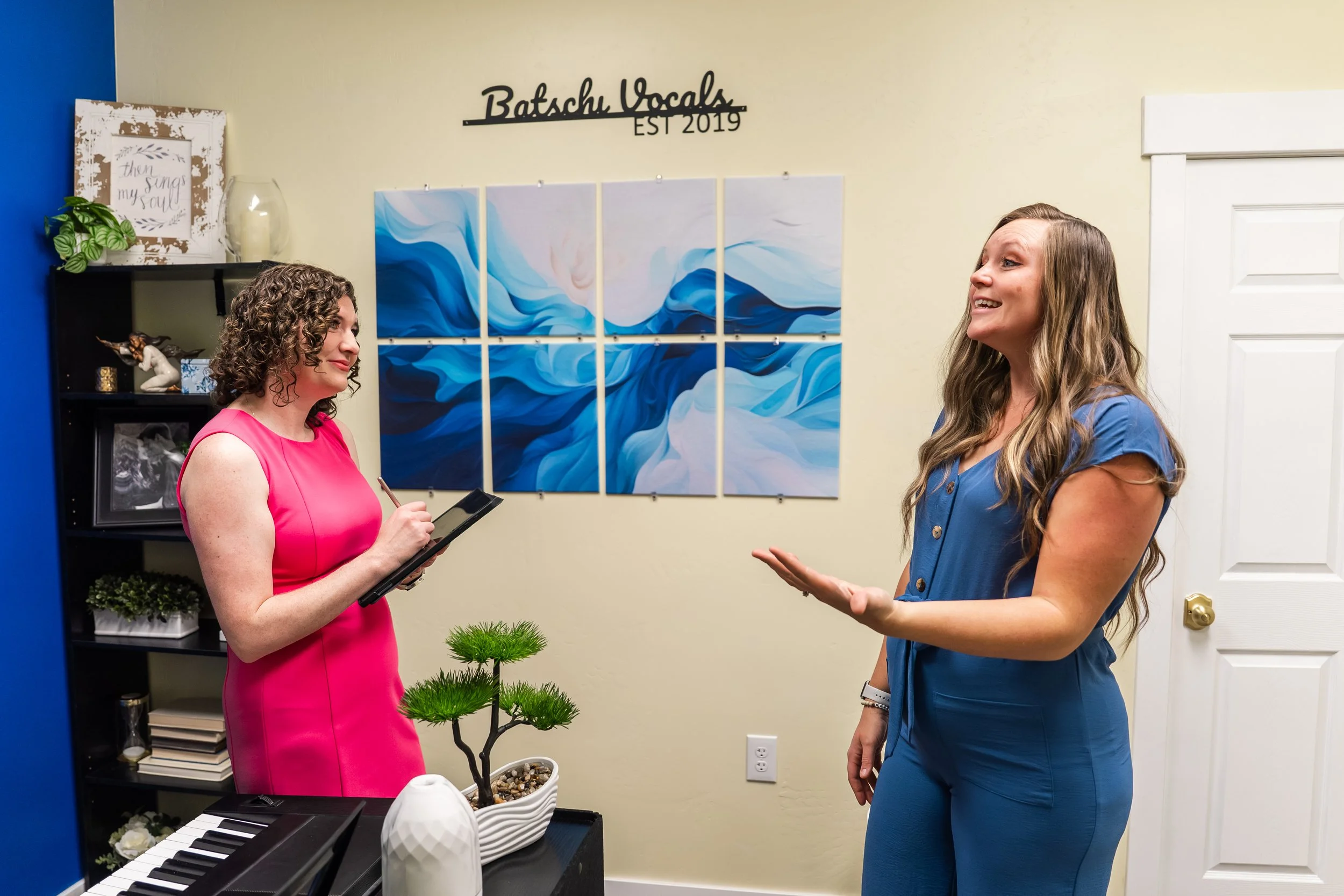 Two women are having a conversation in a room decorated with artwork and a plant. One woman wears a pink dress and holds a clipboard, while the other wears a blue dress and gestures with her hand.