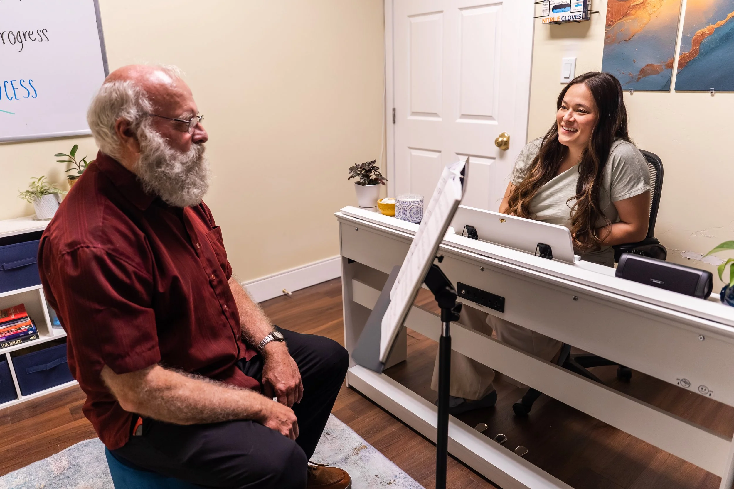 A man with a white beard sitting in a waiting room, smiling and looking at a woman who is sitting at a desk. The woman has long dark hair and is smiling back. The room has a whiteboard with writing on it, a white desk with plants, tissues, and a music stand, and artworks on the beige walls.