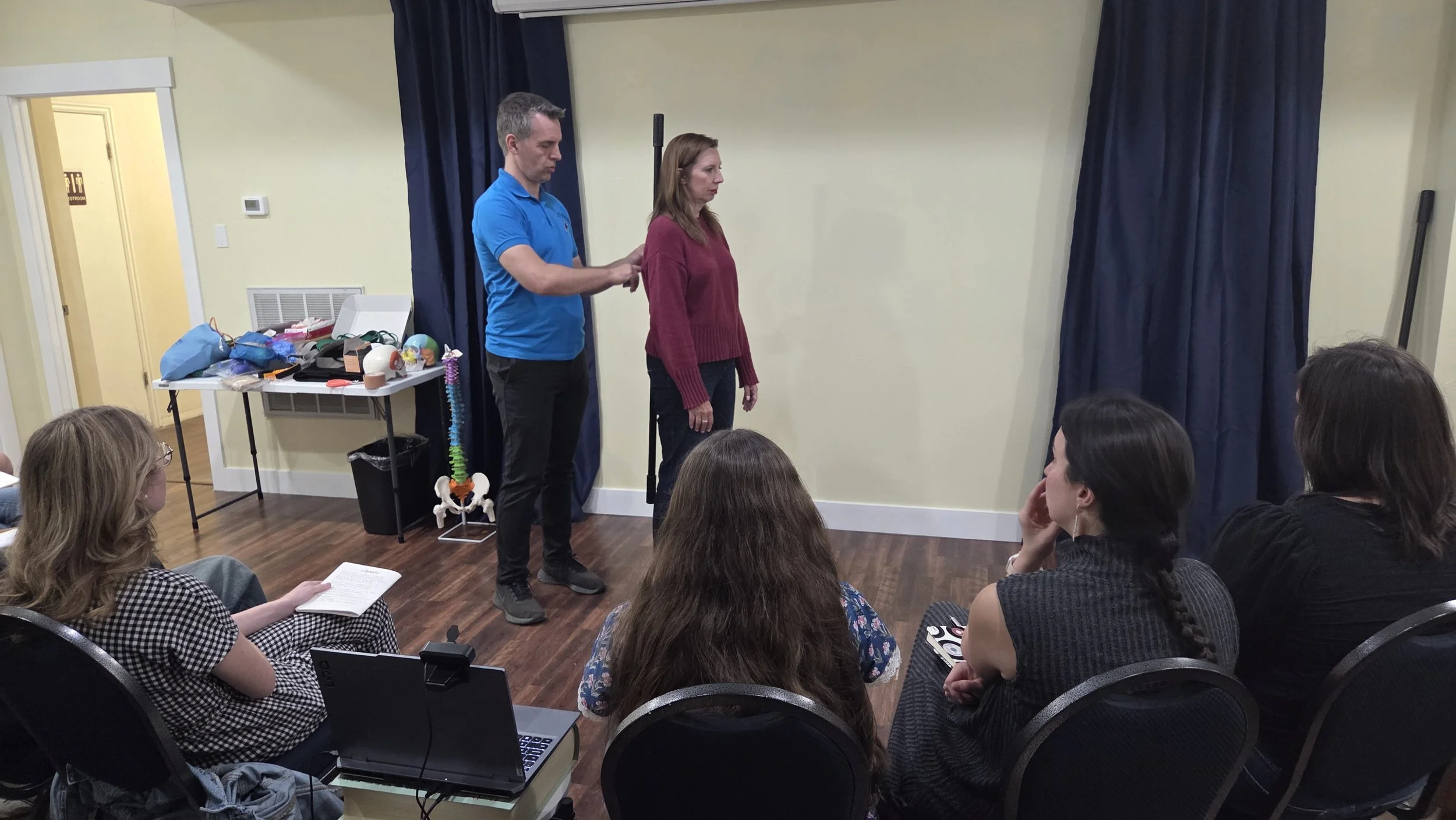 A man in a blue shirt and a woman in a maroon sweater participating in a demonstration or presentation in front of an audience seated in a room. The room has a table with various models and objects, including a spinal model, medical supplies, and a laptop.