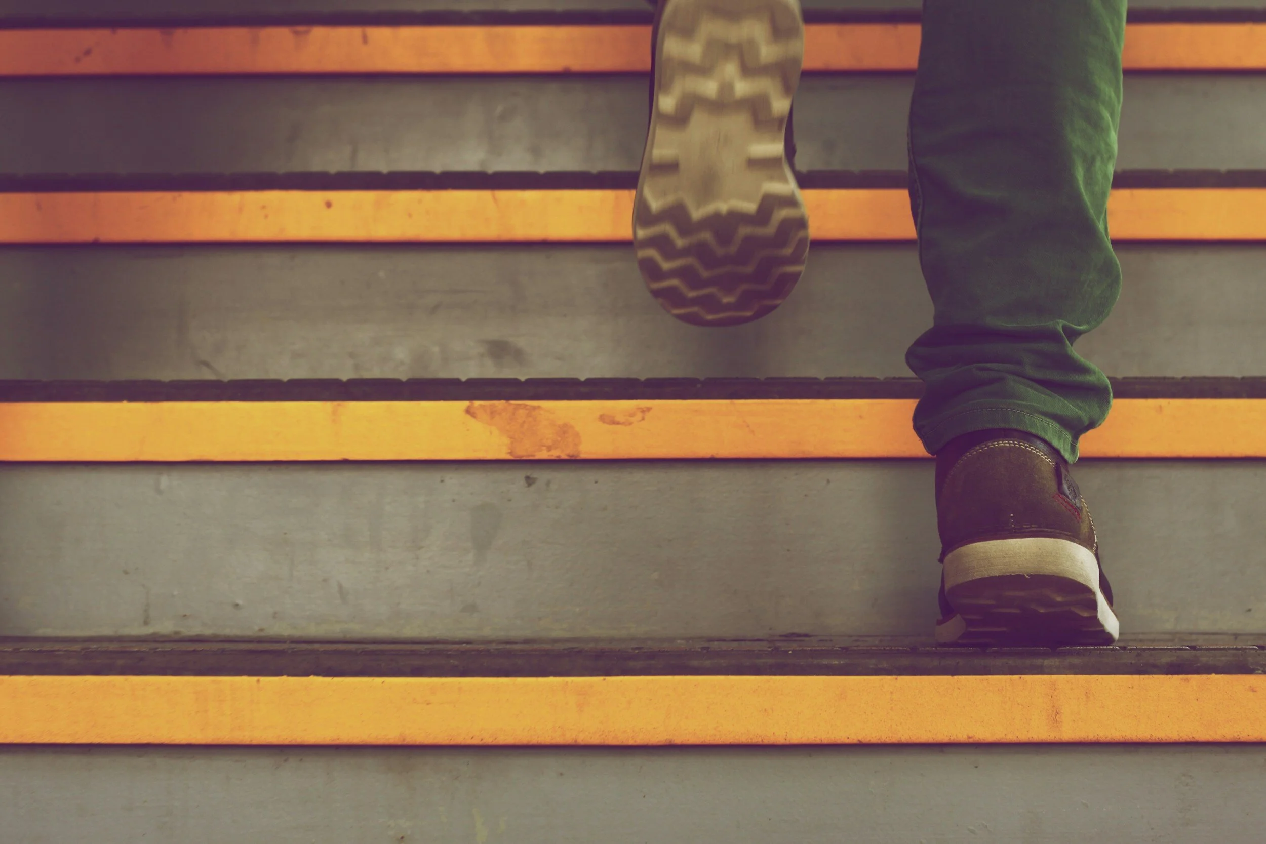 Close-up of a person climbing stairs, with focus on their brown shoe and green pants, as they ascend a staircase with yellow safety strips.