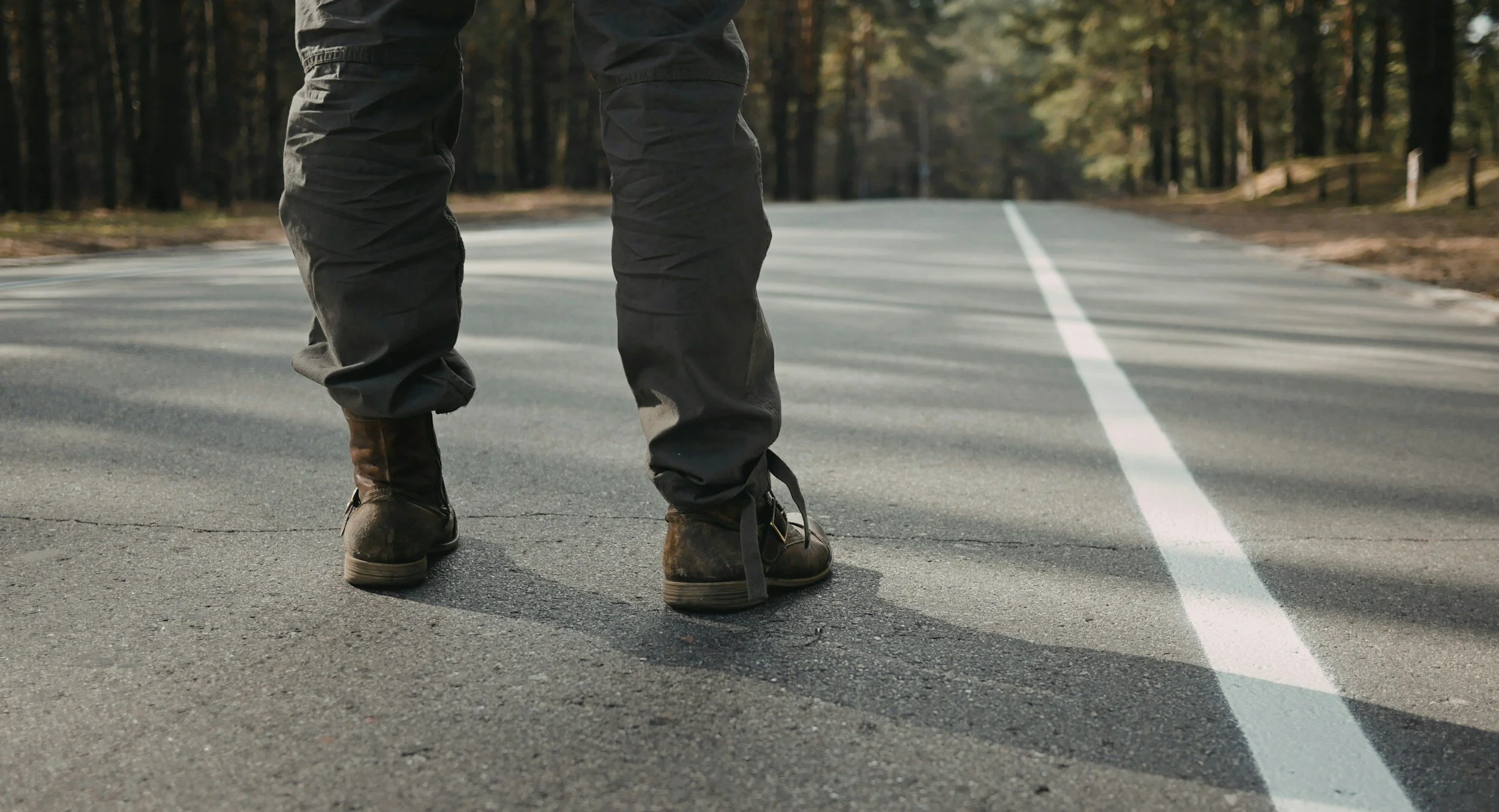 Person standing in the middle of an empty road in a forested area, wearing black cargo pants and rugged brown boots.