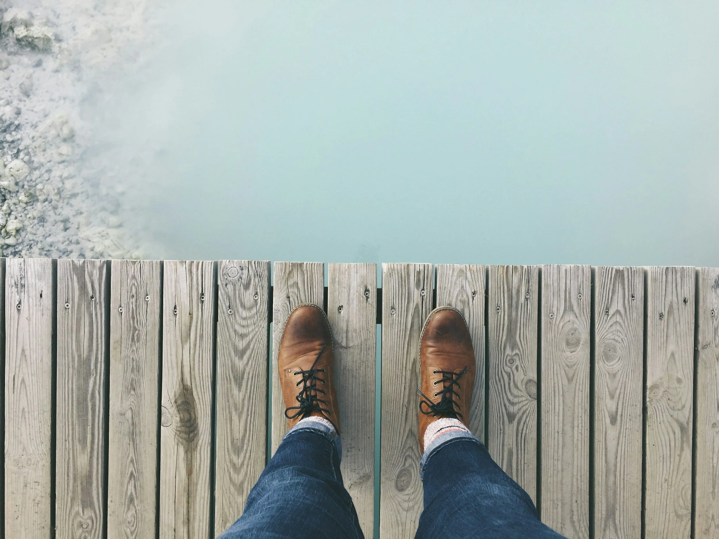 A person wearing brown boots and blue jeans standing on a wooden dock over water.