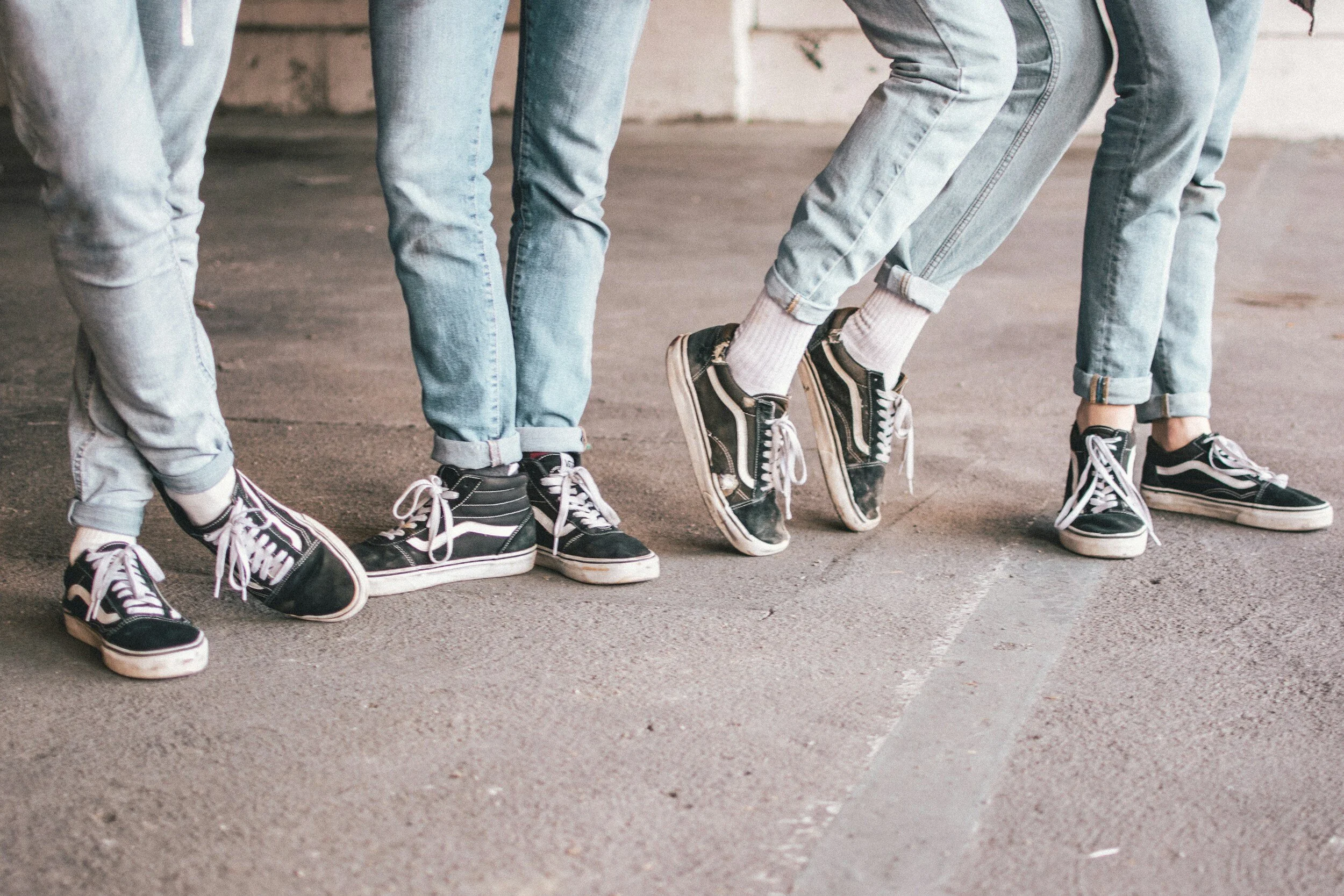 Four people standing close together, all wearing jeans and Vans sneakers. Their legs are visible from the knees down, and they are on a concrete surface.