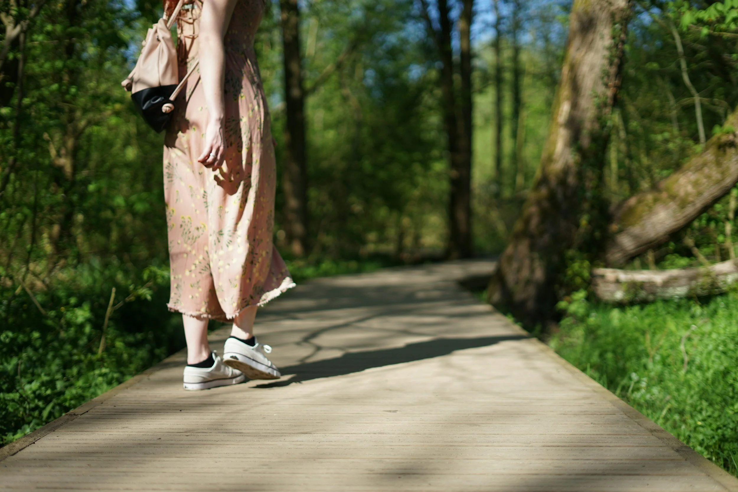 Person walking on a wooden path in a forest, wearing a pink floral dress and white sneakers, carrying a beige backpack.