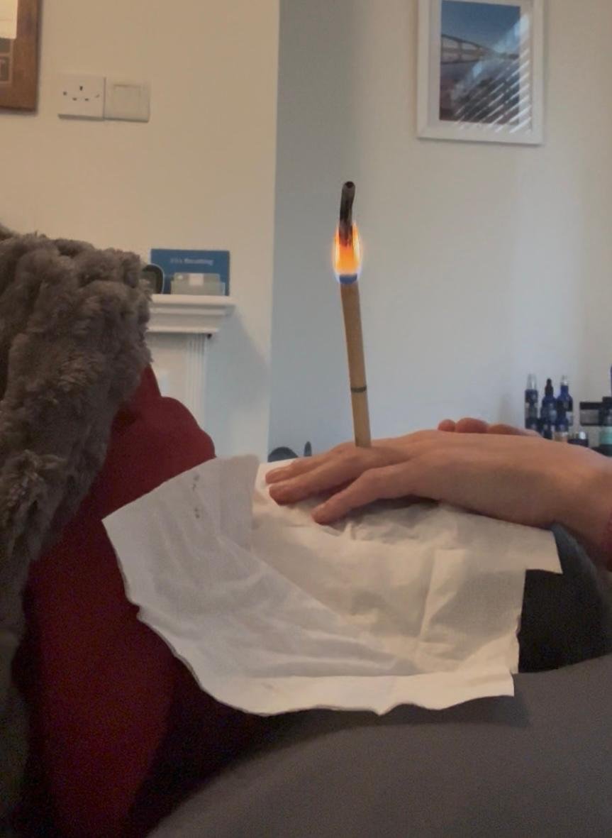 A person is relaxing on a couch with a red pillow and a fur blanket, holding a paper or tissue on their lap, with a burning stick resting on their hand, which has a small flame at the tip.