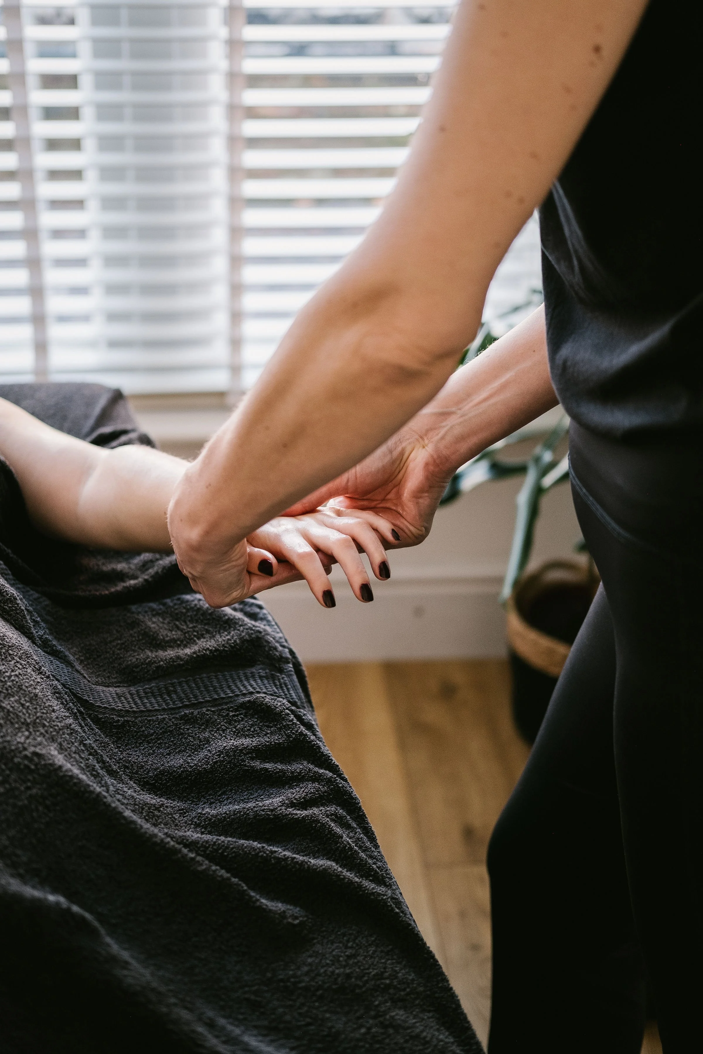 A person receiving a hand massage from a massage therapist in a room with window blinds.