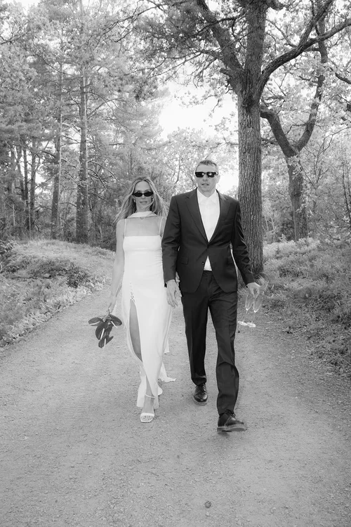 Bride in white halterneck silk dress and groom in black suit both wearing sunglasses walking hand in hand through a pine forest path in the Malaga hills,