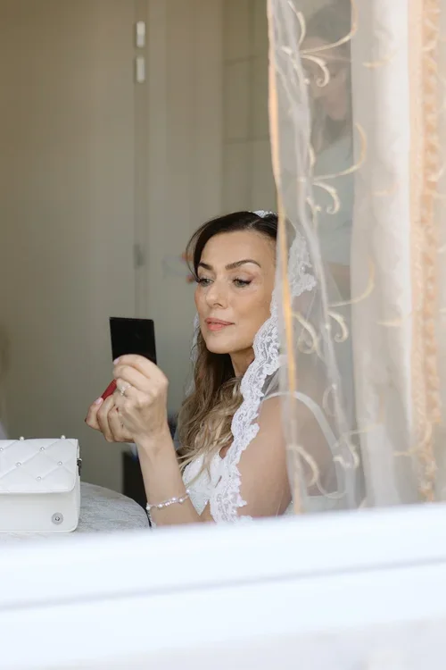 Bride in lace veil checking her make-up in a hand mirror by a sunlit window at a Marbella hotel on her wedding morning