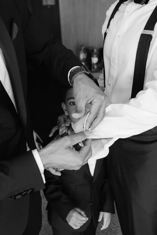 Black and white close-up of father and son buttoning wedding shirt cuffs with a child peeking between them