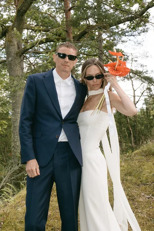 Bride in white halterneck dress holding red anthurium bouquet and groom in navy suit both wearing sunglasses posing together in a woodland setting