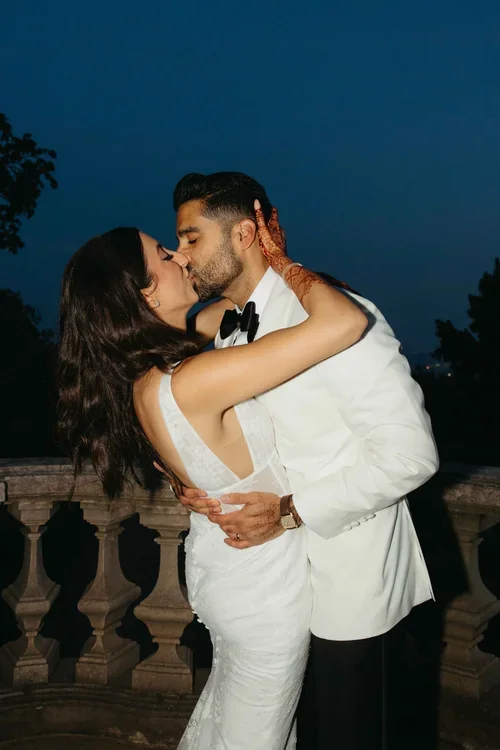 Bride in backless lace wedding dress and groom in white dinner jacket kissing passionately on a stone balustrade terrace under a deep blue night sky
