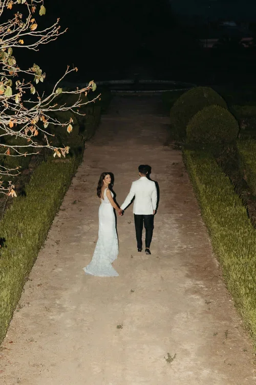 Bride and groom walking hand in hand through a lit manicured garden at night, aerial view destination wedding photography