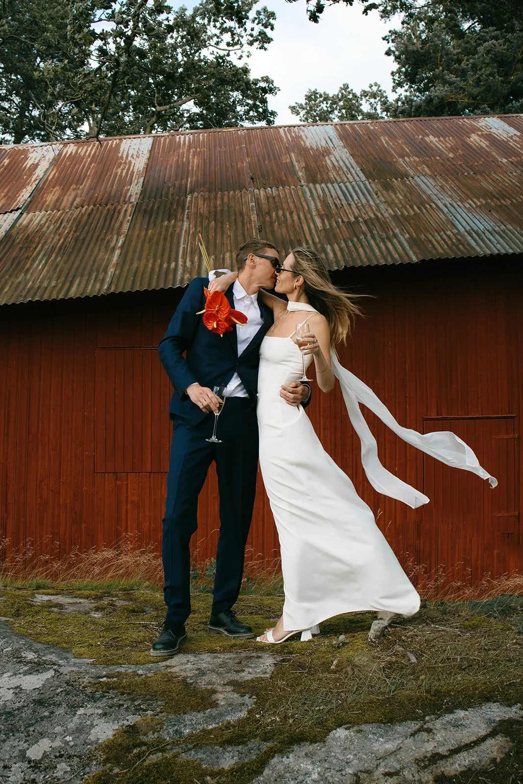 Bride and groom kissing in front of a rustic red barn holding champagne and red anthurium flowers