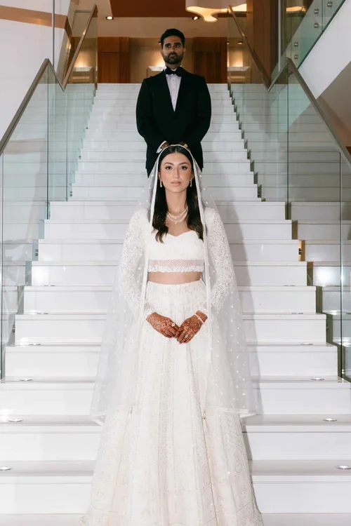 Bride in white embellished lehenga at the base of a luxury hotel staircase with groom in black tuxedo above in Marbella