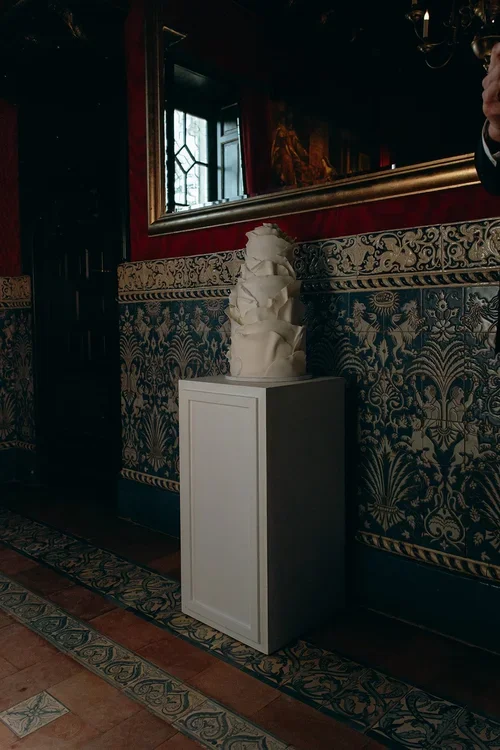 Abstract sculptural white wedding cake on a white plinth against a dark blue decorative tiled wall and gilt mirror inside a luxury castle wedding venue in Spain