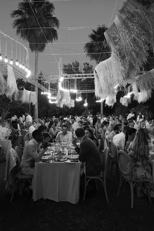 Large outdoor Malaga wedding reception dinner under festoon lights and hanging macrame decorations between palm trees at dusk