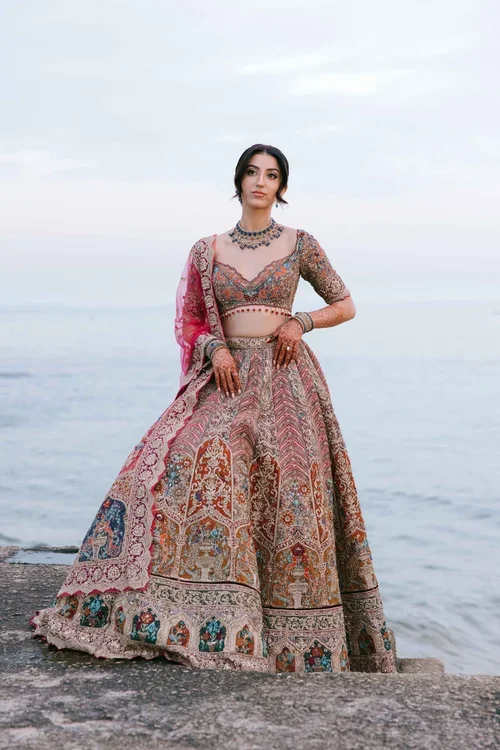 A bride in a embroidered multicoloured lehenga with blue jewellery next to the sea at dusk