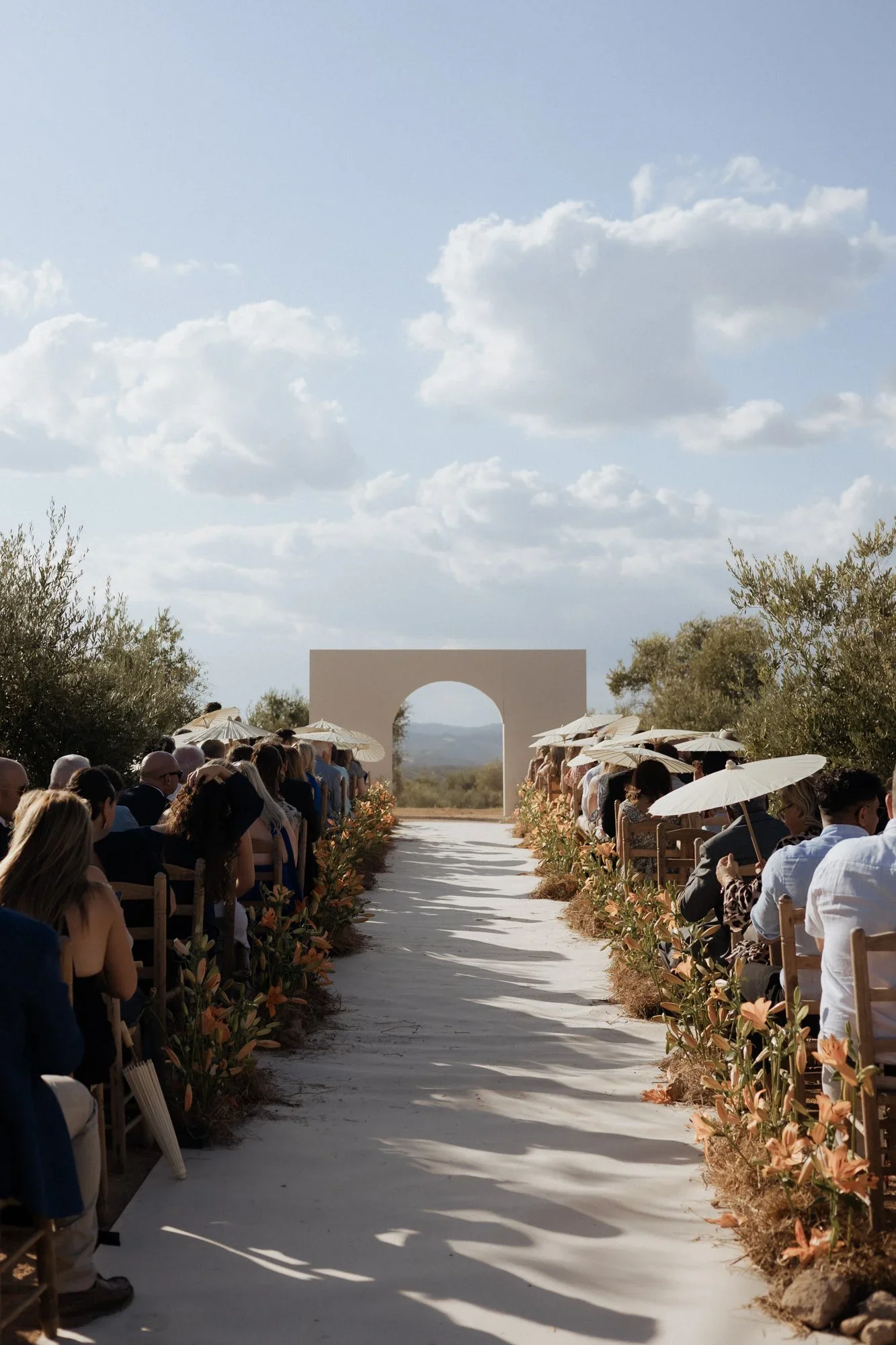 Sun-drenched outdoor wedding ceremony with orange lily-lined aisle, white parasols and a stone arch altar in Malaga