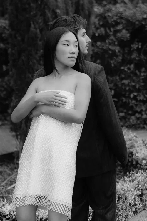 Bride in short white textured mesh dress and groom in dark suit embracing in front of a lush green garden hedge, black and white editorial couple portrait photography Marbella
