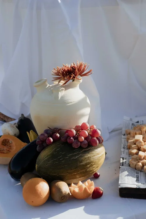 Rustic styled Malaga wedding table display with a white ceramic jug, dried chrysanthemum and seasonal fruit including melon grapes and orange arranged in front of white voile draping