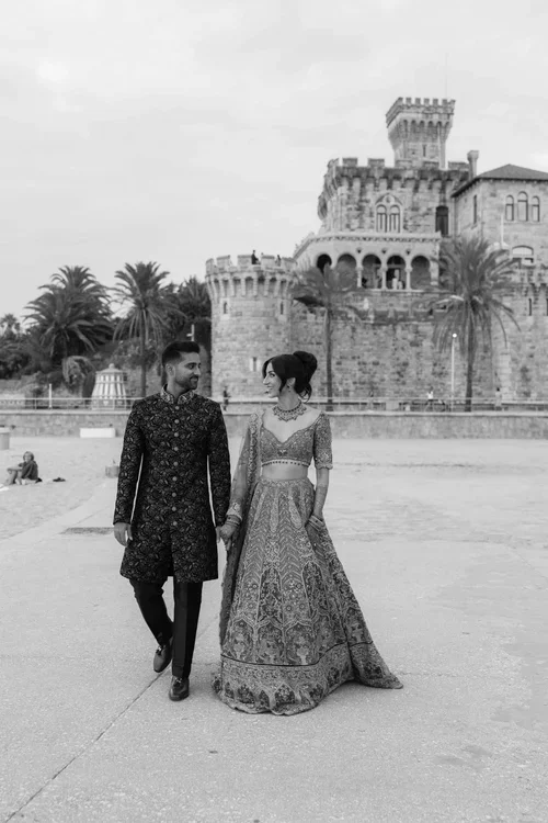 Bride in intricate embroidered lehenga and groom in dark floral sherwani walking hand in hand in front of a coastal castle with palm trees