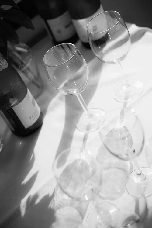 Black and white close-up of wine glasses casting long graphic shadows across a white tablecloth with wine bottles behind