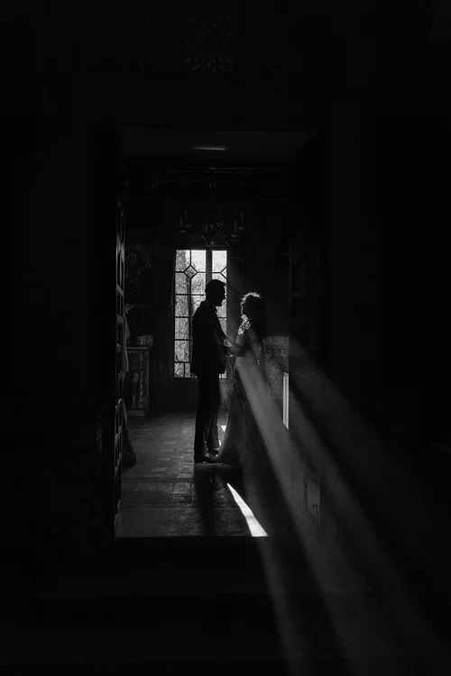 Silhouette of bride and groom standing close together in a dark ornate castle interior with light streaming through a leaded glass window behind them