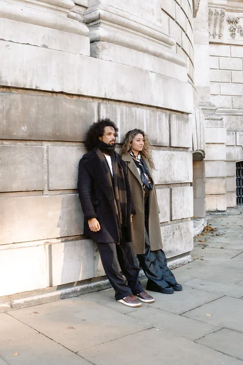 A couple in long winter coats holding hands and leaning on a large stone wall in London