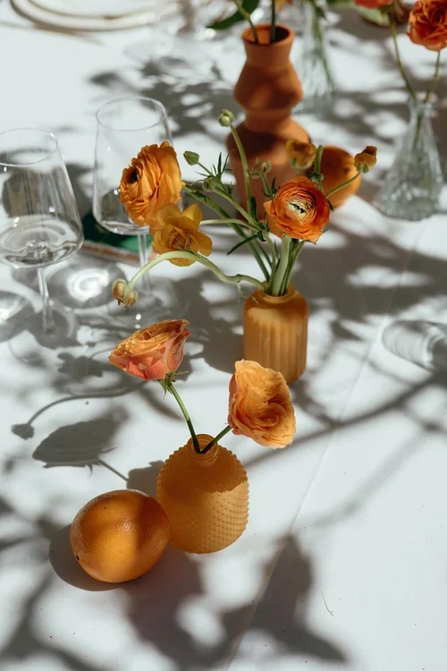 Orange ranunculus flowers in ribbed terracotta ceramic bud vases casting long shadows on a white Marbella wedding reception tablecloth alongside crystal glassware