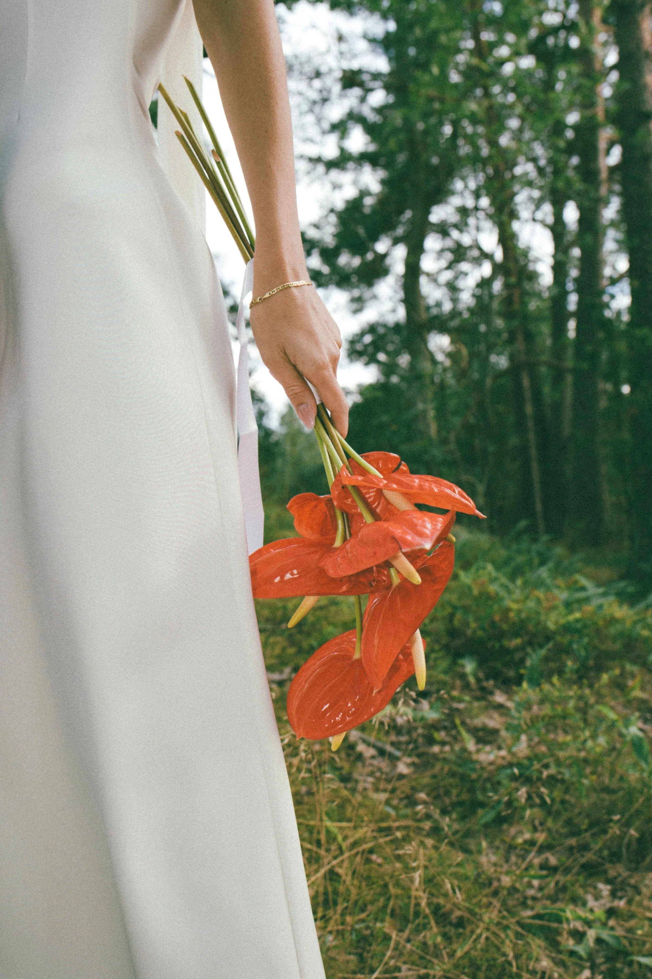 Close-up of bride holding vibrant red anthurium flowers against a white wedding dress with a pine forest backdrop