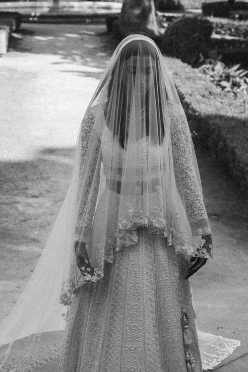 Bride with long lace-edged veil draped over her face walking through a formal garden