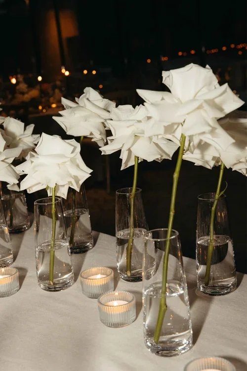 Row of single white roses in tall glass bud vases with tea light candles on a white Marbella wedding reception table at night
