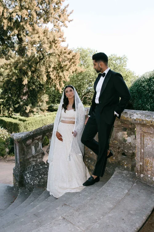 Bride in white embroidered lehenga and groom in black tuxedo on stone steps in a formal garden in Marbella