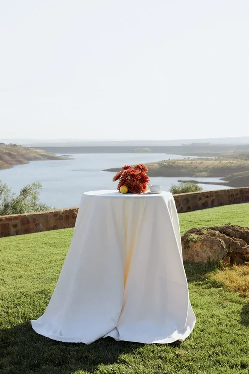 White linen cocktail table with red chrysanthemum floral arrangement and lemon overlooking a sweeping Andalusian reservoir landscape at a Malaga hilltop wedding venue