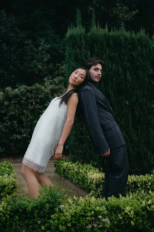 Couple posing back to back in a lush formal garden with tall cypress hedges, bride in embellished white mini dress