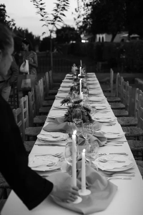 Long outdoor Marbella wedding reception table set for dinner at dusk with tall white candles, mustard linen napkins and dark foliage centrepieces under fairy lights